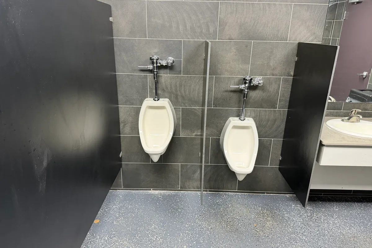 Two white wall-hung urinals with chrome flush valves set on a dark gray tile wall between black privacy partitions in a commercial restroom