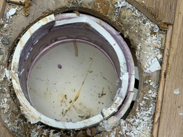 A failed toilet flange seen through a hole sawn in a hardwood floor, its plastic ring cracked into pieces and lifted off the pipe, surrounded by old wax, sawdust, and torn subfloor