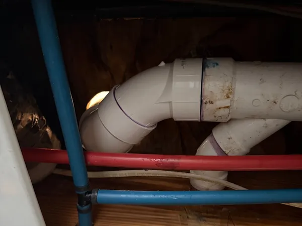 A PVC drain line under a floor turning through two elbows into a tee, with red and blue PEX supply runs and a blue riser crossing the joist bay in front of it