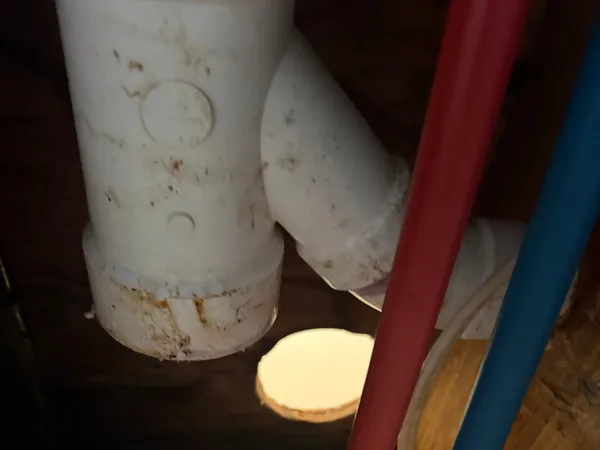 A PVC sanitary tee and elbow on an existing drain line under a floor, stained and dusty, with red and blue PEX supply lines crossing in front of it