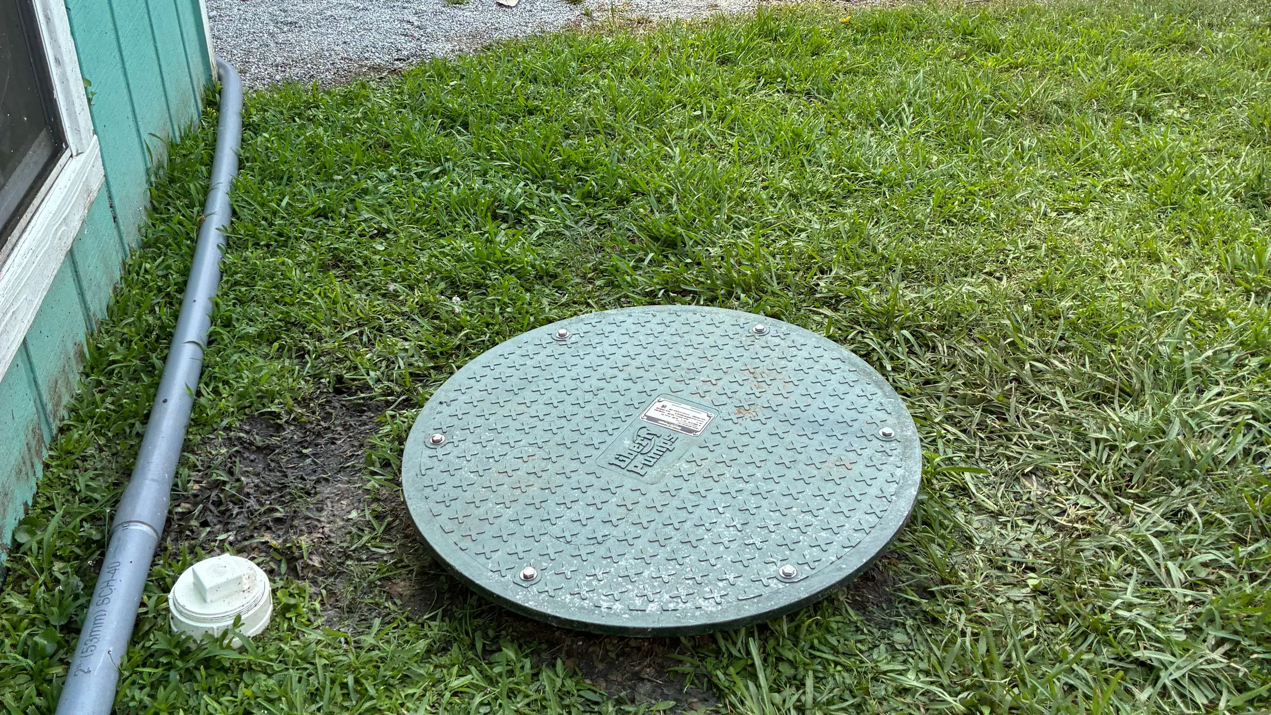 A bolted green fiberglass grinder pump station lid set flush in a mown lawn beside a house, with a gray conduit and a white cleanout running along the foundation