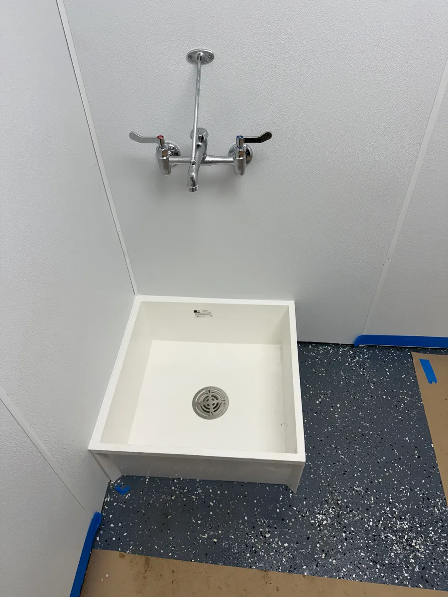 A white floor-mounted mop sink set in the corner of a commercial utility room with a chrome wall-mount service faucet and top brace above it, on a speckled gray epoxy floor