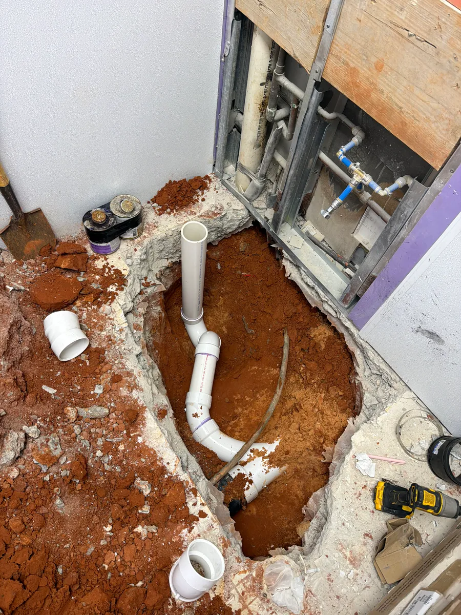 A commercial restroom slab cut open with new white PVC drain line run through the red clay beneath it, elbows and fittings laid out, and the wall opened to expose the existing supply piping