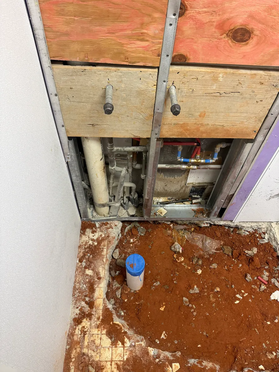 A plumbing chase opened in a commercial restroom wall with copper and PEX supply lines and a PVC vent behind removed plywood panels, and a capped PVC stub-up standing in the excavated slab below