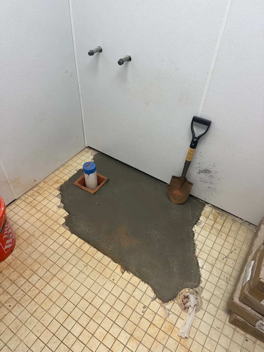A restroom slab patched with fresh concrete around a capped PVC stub-up boxed in wood, two capped supply stubs coming out of the finished wall above it and a shovel leaning beside them