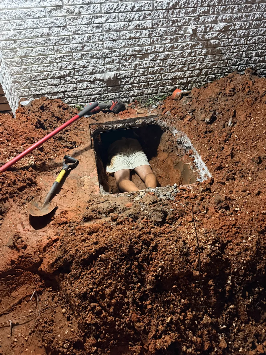 An excavation dug down to a house foundation at night with a square opening cut through the footing wall, a plumber crawling in headfirst, and a shovel and pry bar laid across the spoil pile