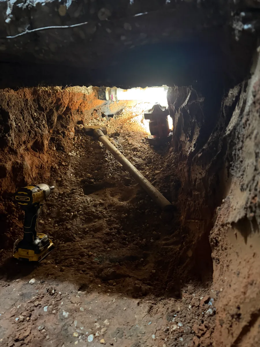 A trench hand dug under a house, the underside of the slab overhead, an old cast iron drain line running down the length of it and a cordless impact driver sitting in the dirt