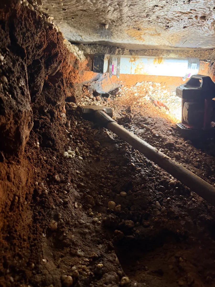 The far end of the tunnel under the house, a new white PVC section of drain line clamped in place tight under the slab with a worklight throwing light down the trench