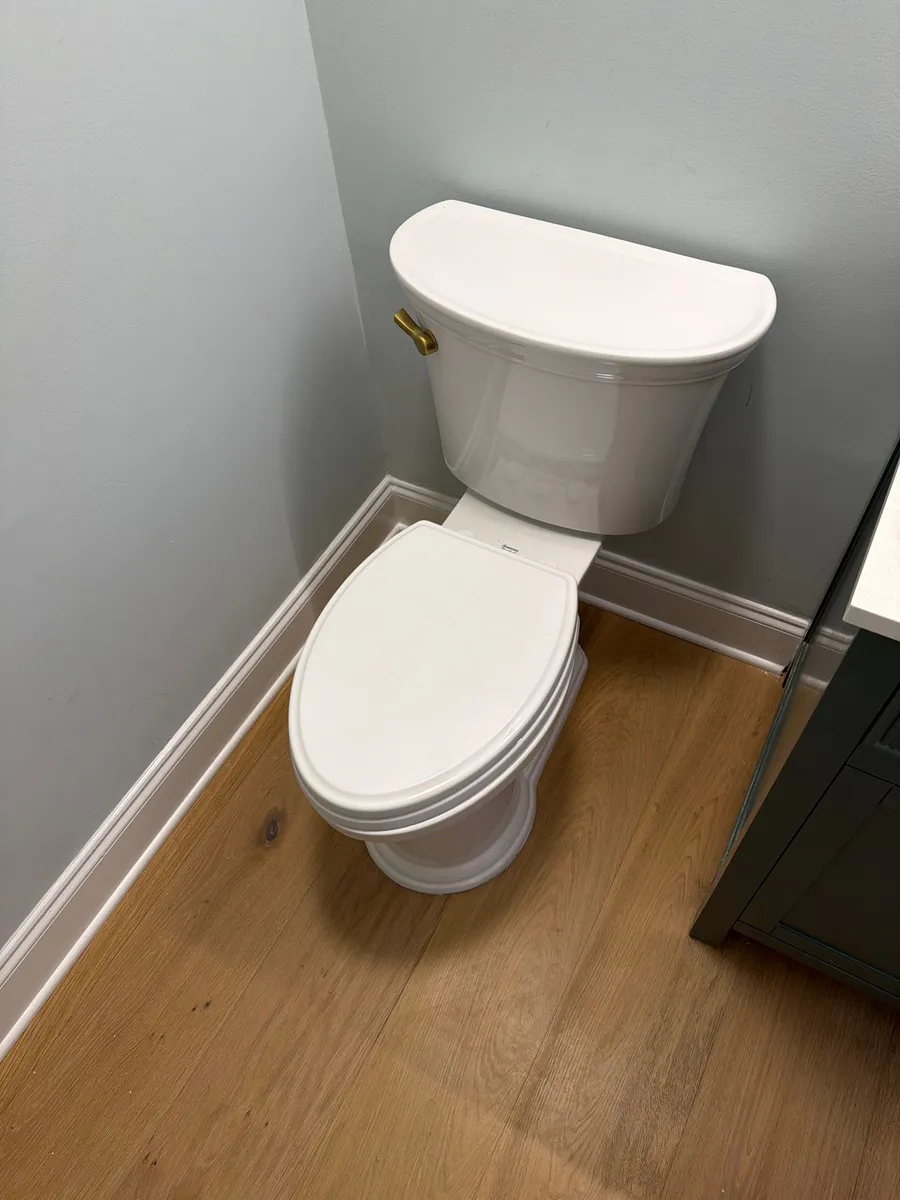 A white two-piece toilet set and trimmed out in the corner of a finished bathroom, seat down, tank against a painted wall above the baseboard, on wood-look plank flooring
