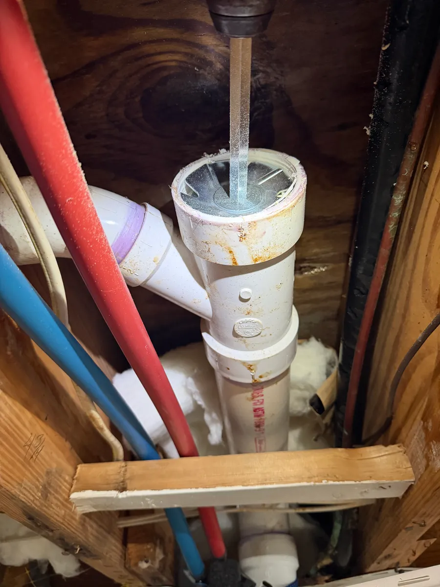 A PVC drain riser and wye fitting standing between floor joists in an open cavity, primed at the joints, with red and blue PEX supply lines running past and batt insulation behind